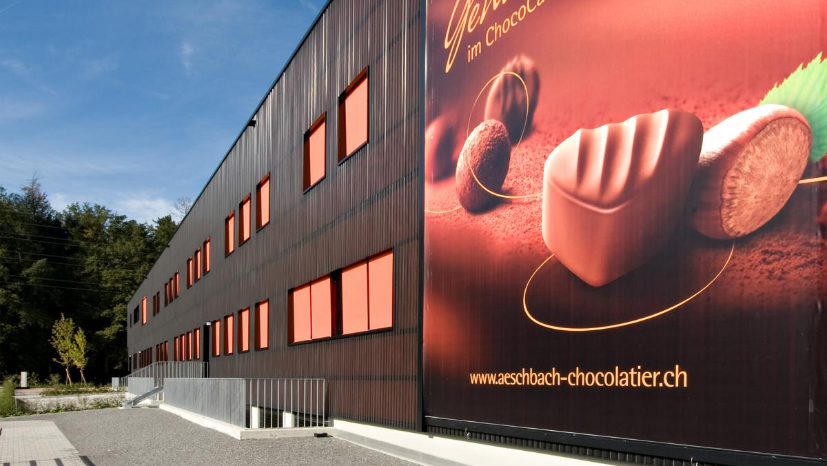 PR Kampagne Chocodromo: Der Firmenhauptsitz von Aeschbach Chocolatier mit einem Werbebanner. Aeschbach Chocolatier verlegt seinen Firmenhauptsitz in den Kanton Luzern und nutzt den Neubau am neuen Standort, um sein Angebot mit neuen Geschäftsbereichen insbesondere im Eventbereich zu erweitern. Diese neuen Entwicklungen werden im Rahmen einer PR-Kampagne kommuniziert.